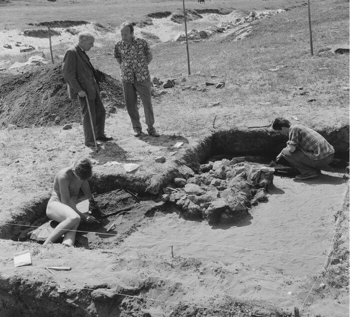 Historisk svart-hvitt bilde av en utgravingsplass hvor to personer sitter i utgravingene og jobber. To personer står ved siden av og ser på arbeidet som utføres.