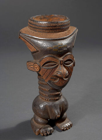 (UEM10192) Drinking vessel carved in the shape of a standing male figure. The vessel is carved in the shape of a man with a decorated, shaved hairline — then a traditional style typical for high-ranking people of the Kuba. The geometric iron pattern on the human-like male figured cup resembles motifs embroidered on Kuba refined raffia textiles. Carved drinking vessels were designed for drinking palm wine. Among the Kuba, most of these cups appear to be sculpted in the form of male figures. It is said that the reason was that the Kuba acknowledged that palm wine was to be drunk by men only. Kuba society is known for its craftsmanship and applied art. Objects of artistic and practical value were appreciated and could signal the owner’s social rank in the community. Like most drinking vessels from the Kuba, this one has no handle. Material: Wood and iron Region: Kasai Occidental, Kuba Height: 22,5 cm. Arrival: Bought by the Museum from B. Brodahl, Trondheim, 1900.