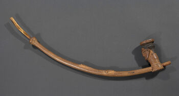 (UEM10201) Wooden tobacco pipe in a human-like form with a bone pipette. The pipe bowl is carved in the shape of a human head with an elaborately carved face and hairstyle. Between the pipe stem and the pipette, a carved geometrical pattern typical of the Kuba is visible. Typical of the Kuba pipes is also that the pipe stem goes upwards in a striking bend. Tobacco smoking was an established practice among the Kuba since the 16th Century. Possibly, the social practice was introduced by the Portuguese, who brought tobacco from the Americas. The ornate pipes are said to have been carved for ceremonial use. Material: Bone, wood Region: Kasai Oriental, Kuba Length: 74 cm. Arrival: Bought by the museum from B. Brodahl, Trondheim, 1900.