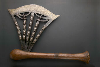 (UEM14570) Ceremonial axe with elaborately forged iron with 42 incised human-like faces or masks. The handle is made from wood covered in copper, decorated with wrapped copper strips in the centre. Crafted by skilled blacksmiths, the axes demonstrate expertise in iron and copper working, a craft closely linked to the spiritual realm. Ceremonial axes symbolise power and authority, serving as a royal emblem. Material: Iron, copper, and wood Region: Kasaï Oriental, Songye Hight/handle: 44,5 cm, width/blade: 34 cm. Arrival: Donated to the Museum by Dr Inge Heiberg, 1905.