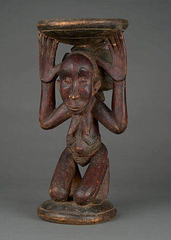 (UEM17775)&amp;#160;Elaborately carved throne and ceremonial seat in the form of a kneeling woman carrying a circular seat on her head. The seat is a receptacle for the chief’s spiritual power, serving to preserve relationships between the ruler, the people, ancestors, and the spiritual world. Sculpted in the form of a female figure, the throne conveys how Luba is a matrilineal society where succession follows the mother’s lineage. A woman supporting the ceremonial seat reveals that although only men are made chiefs, they obtain this position through the mothers’ lineage. Artistically carved, this throne depicts a woman with a sophisticated hairstyle and intricate tattoos on her belly and thighs, signifying a woman of high social rank. The throne is made by a renown woodcarver and Buli master from Kateba. Material: Wood Region: Kasai Oriental, Luba Height: 50 cm. Arrival: Donated by Captain Sundt to the Museum, 1909.