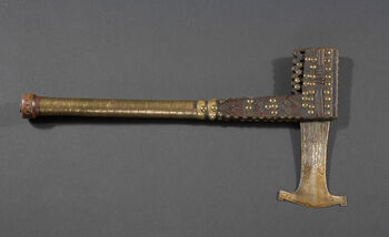 (UEM19218)&amp;#160;Ceremonial axes were prestige items, rather than functional tools. Intricate abstract engravings decorate the iron blade. The lower part of the wooden handle is wrapped in copper strips, while the upper part shows three human-like faces or masks, in between a pattern made of driven brass nails. Ceremonial axes were used by chiefs, diviners, and spiritual leaders to symbolise authority and social rank during ceremonies and important rituals. Elaborate versions of axes were made as royal sceptres and potent symbols of artisanal and aesthetic achievement. Material: Iron, wood, brass, and copper Region: Kasaï Oriental, Chokwe Height: 45 cm, width: 8,8 cm, axe blade: 12 cm. Arrival: Donation from Lieutenant Finn Kjelstrup to the Ethnographic Museum, 1911.
