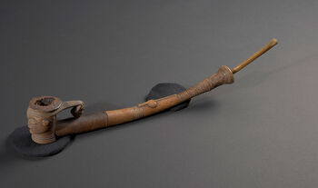 (UEM29478)&amp;#160;Tobacco pipe made in a human-like form. The pipe bowl shows a human head with an elaborately carved face and hairstyle. Necklaces of a geometrical design are engraved around the neck, a design also found on the pipe stem, which goes upwards in a striking bend. The pipette is made of bone. The pipe’s composition shows similarities with ornate tobacco pipes from the Kuba region. Tobacco smoking was an established practice among the Kuba since the 16th Century. Possibly, the social practice was introduced by the Portuguese, who brought tobacco from the Americas. The ornate pipes are said to have been carved for ceremonial use. Material: Bone, iron, and wood Region: Kasai Oriental, Kuba Length: 54,5 cm. Arrival: Donated to the Museum by Johan Cammermeyer, 1923.