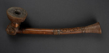 (UEM8171)&amp;#160;Ceremonial tobacco pipe with a protruding bowl decorated with a copper rim and carved in the form of a human head with a delicately designed face or mask. Its straight stem and pipette are neatly decorated with engraved geometrical patterns and copper, and are a distinct element found in some, but not all, Kuba pipes. The Kuba are renowned for their distinctive artwork, including unique pipes with stems carved separately from the bowls themselves. These pipes are not merely utilitarian objects; they are also significant components in ritual and social life, marking prestige and status. Among the Kuba, pipe smoking was a ritual act linked to the invocation of ancestors and mediating to the spiritual world. Ceremonial artefacts reflect the interweaving of society, aesthetics, politics and the sacred world. Material: Wood, metal, and copper Region: Uele and Sankuru, Kuba Length: 41 cm. Arrival: Bought by the Museum from Captain Christian Martini and Captain Christian Schønberg, 1890.