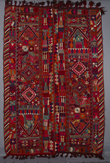 UEM49202. The cloth under the embroidery is somewhat darker than the other blankets in the series. The outer vertical border of each panel has the characteristic multi coloured diagonal lines pattern. In the centre of each panel running vertically are two big diamonds and a mosque or a house at one end. This latter motif is at diagonally opposite ends of the panels. Delimiting the ends of the two panels is a V motif like a roof. Additional motifs consisting of a profusion of living creatures: man, birds, fish, dogs, horses ,insects and so on. These are interspersed with vegetal and geometric motifs. The fringes are plaited. This blanket is especially bright and fresh. 