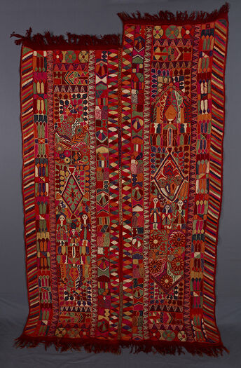 UEM49203. This blanket has double borders on either side of both panels. There is the characteristic diagonal lines pattern on the outer edges and a border of diamonds on the innermost borders of the panels. In the centre of each panel there is a big diamond motif and on both sides of the panels, possibly a roof delimiting the centre field where there are plants and a vase/perfume-like motif. Other motifs consist of geometrical/architectonic motifs, vegetal and living creatures such as female figures in a row of threes, camels, bird atop a tree et cetera.  