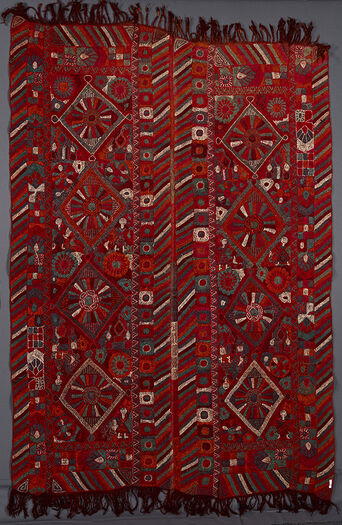 UEM49205. The blanket has double vertical borders.  The characteristic multi coloured diagonal lines pattern are on all of the four vertical edges of the two panels. Each panel has four diamonds in which there is a large flower or a wheel-like motif with rays. There are girls surrounding two of the diamonds in two of their corners and in the other two diamonds in there stylised mudhifs (local guest houses ) in two of the corners. The embroidery is somewhat crude. 