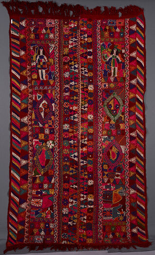 UEM49208. This blanket has double borders. Only the outer edges of the two panels have the characteristic multi coloured diagonal lines pattern. The first vertical panel consist of a chequeredboard of flowers, a girl with long hair wearing a short dress. In centre of the panel is a diamond with flowers, a diamond with a rabbit and two pomegranates, two long legged camels, one which has a rider and chequered board of flowers.    The second vertical panel has a chequered board of flowers, a crowned female figure surrounded by animals and a coffee pot. In the centre of the panel there is a diamond with flowers surrounded by animals, a diamond with a fish surrounded by animals; a big and a small camel with a man holding one of them; chequeredboard of flowers.   The colours are more garish and embroidery rougher than on the other blankets.    The central area of each panel is divided into four sections which are further separated into panels of a floral motif. In the centre are human figures (female figures with short dresses), houses, camels,riders, fish, rabbits, scorpions, birds and multi-petalled flowers as well as four-petalled ones and a pomegranate motif. To be checked and completed. 