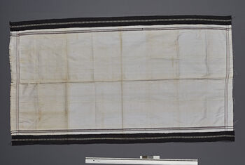 (UEM46903) Sheet. Loin loom, warp faced weave. Woven in two lengths and stitched together. The black side band is woven separately and stitched. Homespun cotton. Pelu is a personal item. During the wedding, the bride gives a Pelu Amar to the groom. He keeps and uses this sheet throughout his life until his death, where it will be used as shroud. During the wedding, a bamboo mat it first put on the ground. The groom’s pelu is then laid on the mat, on the top is the pelu of the bride, and this is laid down before the bridal couple can sit down for the wedding ceremony.