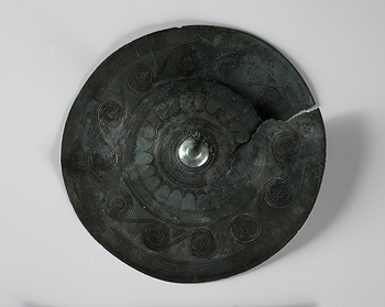 The belt plate from Vestre Evang on Toten in the Inland is one of the finest Bronze Age jewellery we have in the collection. It was found in a large burial mound around 1870. Probably the buried was a woman, who lived once between 1500 and 1300 BC. The belt plate is approx. 13 cm in diameter, and on the back, there is a small loop. The plate is decorated with spirals and sun symbols. The fine and precise lines show that the jewellery was made by a master blacksmith. Belt plates can be compared to today's National costume jewellery, and were worn in the belt on the women's suit. In 1971, almost 3300 years later, the belt plate from Evang was recreated as a jewellery needle for the Toten traditional costume. C5573.