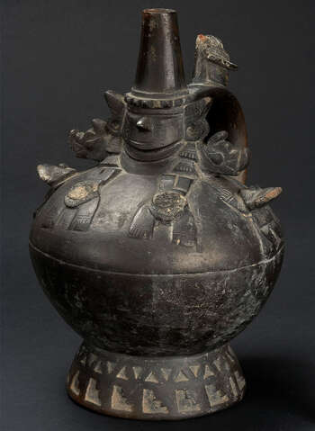 (UEM9970) Globular bottle on a pedestal with excised designs: triangles and steps. The dignitary (“Lambayeque Lord”) with almond shaped eyes, a prominent nose and thin lips, wears a conical headdress, forming the spout and ear ornaments. He is flanked by four human figures lying on the shoulder of the chamber. Mold made. Smudge fired black ware. Middle Sicán, 900–1100, Peru.