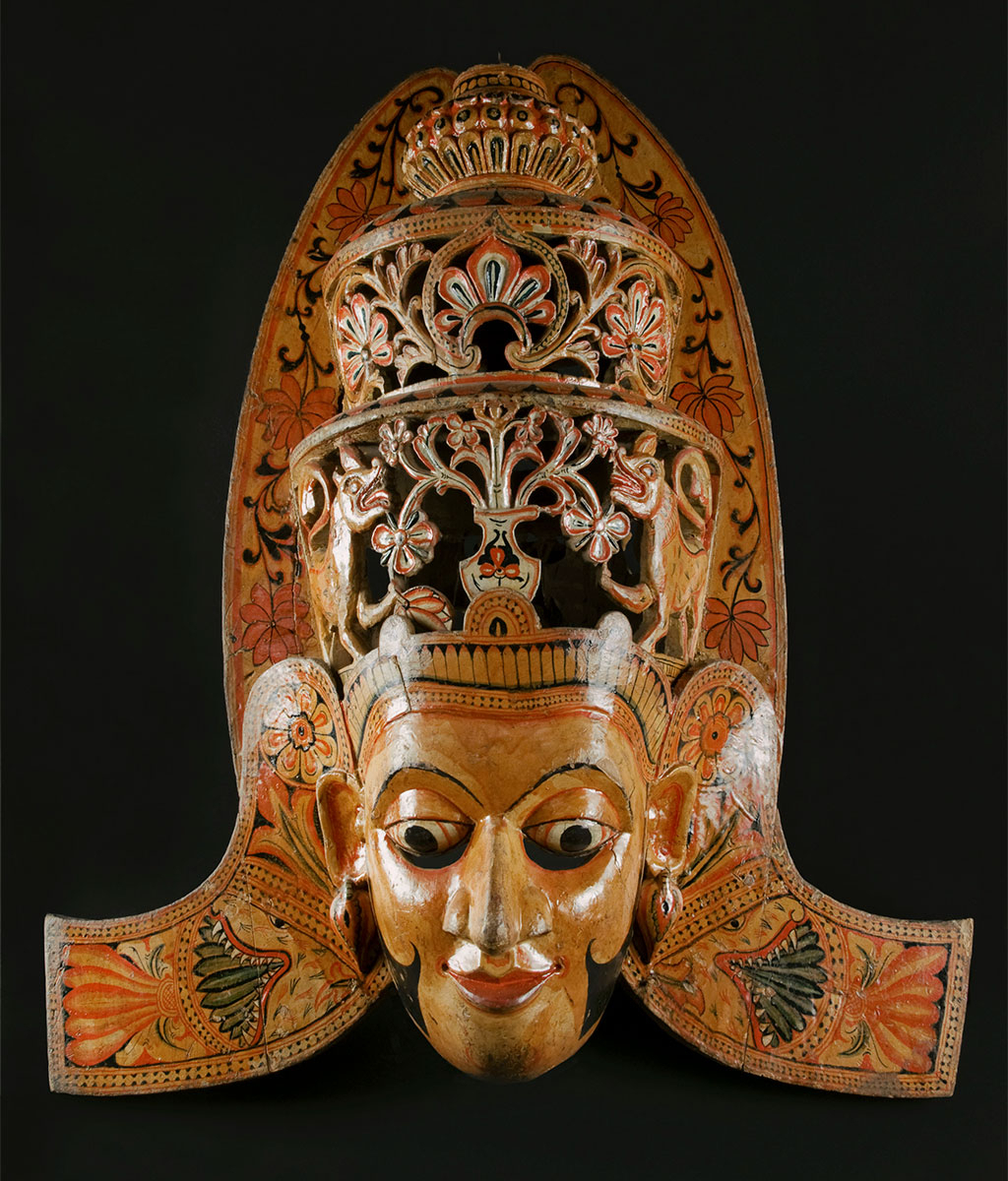 Ritual Function and Context: Kōlam theatre. UEM6999 represents a legendary character of King Mahā-Sammathā performed in the Kōlam theatre. The dancer who wears this mask does not perform with complex dance movements. Instead, the character comes with subtle step movements towards the ritual area ( kōlam madūwa ) with his Queen wife and fellow members and watch the masked dances. The key iconographic features of UEM6999 are: • Pagōda ( stūpā) like crown decorated with intricate Sinhālese ancient motifs ( liyāwelā, palā-pethî, atā-pethî-malā ) • Crown ended with a bud of lotus, the symbol of wisdom ( niřvānā ) or flower called queen of the night ( kadūpul malā ) • Two lions symbolize royalty • Fair skin tone, balance face and long sideburns or moustache • Full head covered mask • Lion heads on each side on the shoulder • Influence of the iconography of Buddhist art in temple • This type of character is the tallest mask in Sinhālese masking culture Note: The animal’s symbols depicted in the crown are different according to the regional tradition. For example, Lion and Horse, and birds’ images can be seen in the crown of this character. Tradition: Southern Dance Tradition/Low-Country Dance Tradition. This tradition is popular in Mātara, Bentōta, Ambalaňgoda in the Southern province and Olabodūwa in Rāiygama Kōralē in Western province of Sri Lanka. Materials and Techniques Polychrome carved wood (white, yellow, red, blue, green, black).
