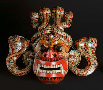 Ritual Function and Context: Kōlam theatre. UEM7000 represents a devil character known as Rathnakūta-Rāksha. This devil mask is associated with a category of supernatural being known as Rāksha. The mask appears in a ritual ceremony known as Kōlam Theatre. In this particular character, the dancer wearing this mask with vibrant costume embodies a very dynamic choreography ( tāndavā-narthanya ) by following the complex drumming notes. This mask can be considered as of the finest examples of this character. The key iconographic features of UEM7000 are: • Red face symbolizes of blood which reflects in Sinhālese name given for this mask as Rathnakūta-Rāksha. • Head-dress and ears (attached separately) formed by coiled cobras. • Protruding eyeballs, widely opened nostrils and mouth with two cobras coming out • Sharp teeth with two long tusks • Painted ancient Sinhālese motifs (mōstarā); leaf motifs (palā-pethî) and fire flames (ginî-dal). • Carving and Painting skills of these decorative elements and tonal qualities of the cobras are based on the skill of mask-carver and tradition. Tradition: Southern Dance Tradition/Low-country dance tradition. This tradition is popular in Mātara, Bentōta, Ambalaňgoda in the Southern province and Olabodūwa in Rāiygama Kōralē in Western province of Sri Lanka. Materials and Techniques: Polychrome carved wood (white, yellow, red, blue, green, black).