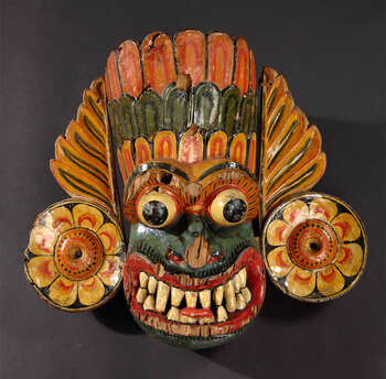 Ritual Function and Context: Kōlam theatre. UEM7002 represents a devil character from Rākshā Cult. The mask appears in a ritual ceremony known as Kōlam theatre ( kōlam natēema ). Two dancers wearing two masks with vibrant costume embody a dynamic choreography ( tāndavā-narthanya ) by following the complex drumming notes. The key iconographic features of UEM7002 are: • Head dress or crown formed with several levels and decorated with Sinhālese motifs ( mōstarā ) shapes of fire flames ( ginî-dal ) or leaf petals ( palā-pethi ). • Circular ear ornaments extend into two flames or foliage. • Although the predominant colour of the face was red, this mask shows a bluish-green tone (discolored) in its face. • Protruding eyes, extended tongues, nostrils turned to the front, and widely opened mouth with sharpen teeth. • Mustache indicated in carving and painting. • Moveable lower jaw. Notes: This character is confused with the character called Marū Rākshā who appears in Kōlam theatre. Tradition: Southern Dance Tradition/Low-Country Dance Tradition. This tradition is popular in Mātara, Bentōta, Ambalaňgoda in the Southern province and Olabodūwa in Rāiygama Kōralē in Western province of Sri Lanka. Materials and Techniques: Polychrome carved wood (white, yellow, red, blue , green, black).