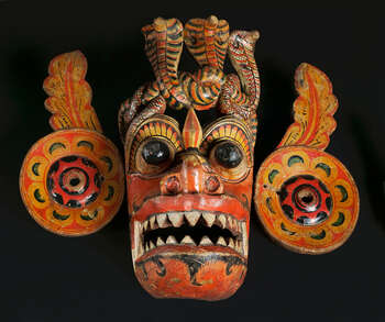 Ritual Function and Context: Kōlam theatre. UEM7004 represents a devil character from Rākshā cult. The character could embody either Nāga Rākshā or Ratnakūtā Rākshā. This mask appears in Kōlam Theatre. The dancer wearing this mask with vibrant costume embodies a very dynamic choreography ( tāndavā-narthānya ) by following the complex drumming notes. The key iconographic features of UEM7004 are: • The head-dress formed with coiled cobras (one is broken) features of Naga Rākshā and Ratnakūtā Rākshā mask. • Face in red colour. • The ears are formed by two discs and extended foliage, similar to the mask of Pūrnakā-Rākshā. • Protruding eyes, extended tongues, nostrils turned to the front, and widely opened mouth with sharpen teeth. • indication of mustache in carving or painting. • Application of traditional Sinhālese motifs ( ginî-dal, palā-pethî, atā-pethî-malā ) Tradition: Southern Dance Tradition/Low-Country Dance Tradition. This tradition is popular in Mātara, Bentōta, Ambalaňgoda in the Southern province and Olabodūwa in Rāiygama Kōralē in Western province of Sri Lanka. Materials and Techniques: Polychrome carved wood (white, yellow, red, blue, green, black).
