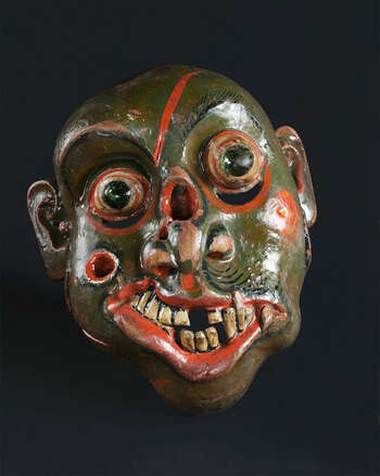 Ritual Function and Context: Kōlam theatre. UEM7011 represents a character of soldier ( hēvāyā ). This character is used in Kōlam Theatre. In Kōlam theare, several hēvāyā masks appear as a group of soldiers. The performance is known as Hēvā-Kōlama. The key iconographic features of UEM7011 are: • The most expressive characters from Kōlam masks. • Deformed facial forms: unfocused ball eyes, marks of wounds, and swollen bumps in the face and mouth, broken teeth. • Leeches coming out of wounds. • Green skin colour makes this mask a rare piece. Mask often comes in deep yellow, orange or white (pale)and rarely in green skin tone. • The traditional mask carvers demonstrate their creative freedom, imagination and skill in carving this character. Notes: These features have made Hēvāyā character identified as a Sanni mask from Sanniyäkuma ritual. Tradition: Kōlam Theatre/the Southern Dance Tradition/Low-Country Dance Tradition. This tradition is popular in Mātara, Bentōta, Ambalaňgoda in the Southern province and Olabodūwa in Rāiygama Kōralē in Western province of Sri Lanka. Materials and Techniques: Polychrome carved wood (white, red, green, black), Glass Balls in the eyes.