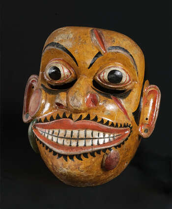Ritual Function and Context: Kōlam theatre. UEM7012 represents a character of Soldier ( hēvāyā ). This character is used in Kōlam Theatre. In Kōlam theare, several hēvāyā masks appear as a group of soldiers. The performance is known as Hēvā-Kōlama. The key iconographic features of UEM7012 are: • The most expressive characters from Kōlam masks. • Deformed facial forms: unfocused ball eyes, marks of wounds, and swollen bumps in the face and mouth. • Leeches coming out of wounds. • Mask comes in deep yellow, orange or white (pale) and rarely in green skin tone. • These features make Hēvāyā character identified as Sanni mask of Sanniyäkuma ritual. • The traditional mask carvers demonstrate their creative freedom, imagination and skill in carving this character. For example, teeth are not represented as broken in this mask. Notes: These features have made Hēvāyā character identified as a Sanni mask from Sanniyäkuma ritual. Tradition: Southern Dance Tradition/Low-Country Dance Tradition. This tradition is popular in Mātara, Bentōta, Ambalaňgoda in the Southern province and Olabodūwa in Rāiygama Kōralē in Western province of Sri Lanka. Materials and Techniques: Polychrome carved wood (white, yellow, red, green, black).