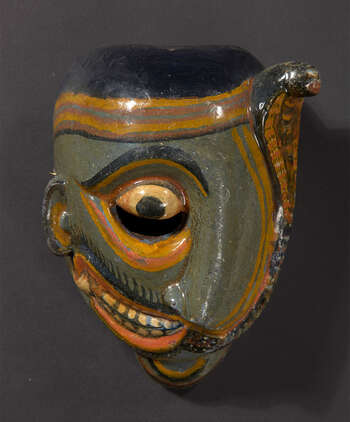 Ritual Function and Context: Sanniyäkuma / Dāha-atā sanniya. UEM7014 represents one of the eighteen masks performed in Sanniyäkuma Ritual. This mask represents blindness ( kanā sanniya ). The dancer wore this mask steps rhythmically into the dancing floor ( sanni vîdiyā ) with aid of wooden stick representing the idea of white cane. Kanā Sanniya creates series of conversation with the main drummer that make the sick person and audience laugh. The key iconographic features of UEM7014 are: • The iconographic features are different based on the reginal traditions. • Covering left eye/ face from a Nāga coming from a nostril or near mouth • UEM7014 is painted neutral green. This mask also comes in vivid red tone. • Smaller in size Notes: Complexity of the iconographic features of this mask: If the nāga covers the right eye of the mask, that mask could represent the deafness ( bîhirî ). This character is also represented with the eyes without pupils and painted in white. Tradition: The Southern Dance Tradition/Low-Country Dance Tradition. This healing ritual is believed to be originated from Bali ritual and is popular in Mātara, Gāll, Bentōta, in Southern province and Olabodūwa in Rāiygama Kōralē in Western province in Sri Lanka. Significant difference can be seen in this ritual held according to Mātara Tradition and Bentōta Tradition. Materials and Techniques: Polychrome carved wood (white, yellow, red, black).