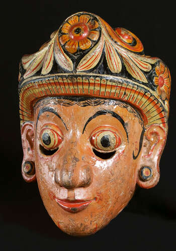 Ritual Function and Context: Kōlam theatre. UEM7024 represents a legendary character of queen. The mask is known as Bisawā or Manāme-Bisawā. The mask appears in Kōlam Theatre. As the mask of Bisawā, this character performs with the King Maha- Sammatha. As the character of Manāmē Bisawa, it narrates the story known as Manāme Kōlama and a story from Jātaka Pothā called Chuĺlā-Danuďdarā Jātakaya. In Sinhālese rituals, all the female characters represented from masks are worn by male dancers. The key iconographic features of UEM7024 are: • Symmetrical and sharply carved facial features ( vatākuru muhunā ) with half-smile ( adavaň sinahawa ) and half circle eyebrows. • Fair skin tone • The crown decorated in Sinhālese floral motifs of eight petals flower ( atā-pethî malā ) and leaf petals ( palā-pethî ) • This kind of low-high floral headdress is typical of the tradition known as Mātara. Tradition: Southern Dance Tradition/Low-Country Dance Tradition. This tradition is popular in Mātara, Bentōta, Ambalaňgoda in the Southern province and Olabodūwa in Rāiygama Kōralē in Western province of Sri Lanka. Materials and Techniques: Polychrome carved wood (white, yellow, red, black).