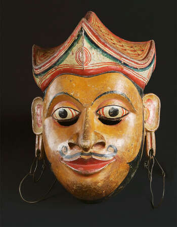 Ritual Function and Context: Kōlam theatre. UEM7028 represents a legendary character. The character is known as Prince/King of Manāmē ( manāmē kumārayā or manāmē rajū ). The mask appears in Kōlam Theatre. The mask plays one of the key roles in the Story of Manāmē or Chuĺlā Dhanūďdarā jātakaya. This character performs in collaboration with the character of Manāmē Bisāwā/Kumāriyā and King of the Ancestor/ hunter ( vadî-rajā ). The key iconographic features of UEM7028 are: • Pleasing facial features of a young man. • Fair skin tone • Curved mustache and sideburns. • The headdress forms similar to the traditional cap of Sinhālese Kings ( nilāmē thoppiya ). (This can also be different based on regional tradition). • Royal signs of Elongated ears (an emphasis) and attached earrings. Tradition: Southern Dance Tradition/Low-Country Dance Tradition. This tradition is popular in Mātara, Bentōta, Ambalaňgoda in the Southern province and Olabodūwa in Rāiygama Kōralē in Western province of Sri Lanka. Materials and Techniques: Polychrome carved wood (white, yellow, red, black), earrings attached separately.