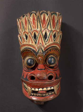 Ritual Function and Context: Kōlam theatre. UEM7038 represents a mythical devil character from Rākshā cult. The mask is known as Puřnakā Rākshā. This mask appears in Kōlam theatre. In some traditions, two masked dancers of this character perform in Kōlam theatre. The performance shows very rigorous movements of dance followed by the drumming beat and singing verses of Kōlam ( koalm kavi ). UEM7038 has lost its ear forms that should be similar to UEM7002 and UEM7003. The key iconographic features of UEM7038 are: • Head-dress formed with several levels • Head-dress decorated in shapes of Sinhālese motifs of flames ( ginî-dal ). • Missing ear ornaments should be like UEM7002 or UEM7003. • Predominated Red in the face. • Protruding eyes, extended tongues and widely opened mouth with sharpen teeth • Indication of mustache in forms of flames • The gaze of the eyes called the gaze of the death ( marū-baĺmā or diśitiya ) • Damaged lower mouth is not movable like some other masks have in general. Notes: This character is confused with the character called Marū Rākshā who appears in Kōlam theatre. Tradition: Southern Dance Tradition/Low-Country Dance Tradition. This tradition is popular in Mātara, Bentōta, Ambalaňgoda in the Southern province and Olabodūwa in Rāiygama Kōralē in Western province of Sri Lanka. Materials and Techniques: Polychrome carved wood (white, yellow, red, black, blue).