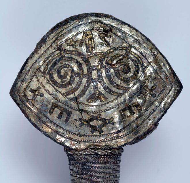 Close-up photo of the pommel. Inlays of gold wire makes up the central line in each symbol. The gold is framed by a copper wire that is now black. All the intermediate surfaces have been filled in with parallel silver wires. At the top a hand holding a cross is visible. Photo: Vegard Vike, Museum of Cultural History, UiO.