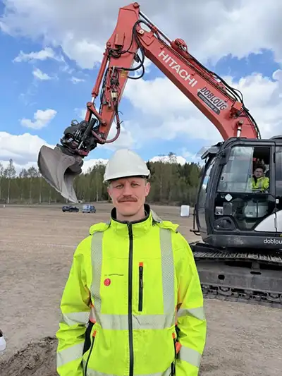 Mann står på et jorde og ser i kamera. Han har på verneutstyr, en gravemaskin i bakgrunnen.