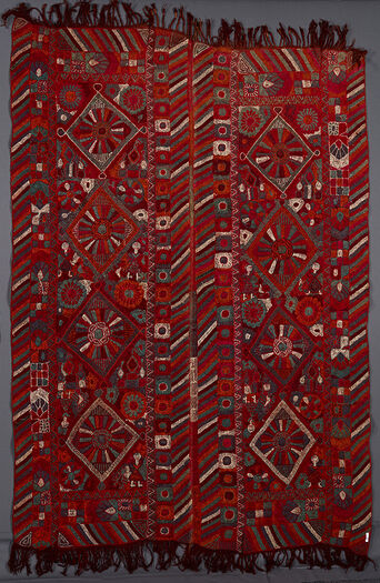 UEM49205. Teppet har doble loddrette border.  Det karakteristiske flerfargede diagonale linjemønsteret finnes på alle fire loddrette kanter på teppets to paneler. Hvert panel har fire diamanter hvor det er en stor blomst eller et hjul-lignende motiv med stråler. To av diamantene er omringet av jenter i to av hjørnene, mens de andre to diamantene har stiliserte mudhifs (gjestehus) i hjørnene.  Broderiet er noe grovt gjennomført.