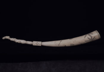 (UEM22753) Utskåret, dekorert elfenben laget av elefantstøttann. Tuppen er skåret ut i form av en tredimensjonal, kvinnelig figur. Støttannens midtre del er dekorert med geometriske og sirkulære utskjæringer, og menneskelignende kvinnelige og mannlige figurer som deltar i daglige aktiviteter, eksempelvis menn som klatrer med tau i palmetrær og kvinner som hugger palmetrær med økser. Disse scenene er skåret i profil. De delikate formene antyder en bemerkelsesverdig håndverks- og kunstnertradisjon. På slutten av 1880-tallet og 1900-tallet var elfenben en viktig vare i regionen. Handelsmenn lagret og distribuerte elfenben, så vel som slaver. Dekorert elfenben viser til en etablert kunstform for gravering og utskjæring av ulike motiver fra daglig- og rituelt liv. Prosessen består av å gravere inn motivet på overflaten, før linjene mørknes av pigment. Teknikken ble også brukes til dekorering av bruksgjenstander, som hårnåler, armbånd, knivhåndtak og til utskjæringer av hele støttenner, selv om sistnevnte er av de mer sjelden blant Mangbetu. Materiale: Elfenben Region: Haut-Uele, Mangbetu Lengde: 59 cm. Ankomst: Donert til museet av advokat Jacob Vogt, 1915.