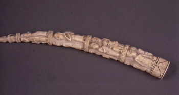 (UEM7398) Utskåret elfenben laget av en flodhest støttann. Tuppen av tannen er skåret ut i form av en knelende kvinnelig figur. Tannens midtre er dekorert med utskårne  motiver av dyre- og menneskefigurer, i all hovedsak kvinner, menn og barn som utfører sine dagligdagse aktiviteter. De mest sentrale motivene på elfenbensskulpturer fra Luango er menneske- og dyrefigurer som inngår i scener fra daglig- og rituelt liv. Figurene er alltid sett fra siden, i profil, mens figuren på tannspissen er tredimensjonal. Blant Luango var det spesialiserte håndverkere som skar ut elfenbensskulpturer. Håndverkerne var høyt verdsatt for sine ferdigheter i faget som ble ansette å være en videreføring av game Luago tradisjoner. Luango-området, hvor elfenbentannen opprinnelig ble utskåret og dekorert, var på slutten av 1800-tallet et blomstrende handelsknutepunkt for europeere og handelsforetak, hovedsakelig fra Portugal og Nederland, men også fra Skandinavia og Midtøsten. Handelen sentrerte seg om elfenben, tekstiler og slaver fra innlandet. Det transnasjonale miljøet fra denne historiske perioden ser ut til å ha påvirket billedspråket til skulptører og andre kunsthåndverkere i regionen. Utskjæringen på denne flodhest-tannen, som viser motiver med kvinner, menn og barn i sine lokale omgivelser, antyder at den kunstferdige tannen antagelig var ment som suvenir for utenlandske handelsmenn som besøkte regionen. Dekorert elfenben av elefant- og flodhest-støttenner ble i utgangspunktet laget for lokal bruk, da som eksklusive artefakter for bruk i seremonier, på kongelige gravplasser, som gaver i diplomatiske utvekslinger, men ble etter hvert også laget på bestilling fra europeere. Materiale: Elfenben Region: Ekvatorial-Kongo, Luango Lengde: 30 cm. Ankomst: Kjøpt av museet fra J. F. G. Umlauff, Umlauff-museet, Hamburg, 1888.