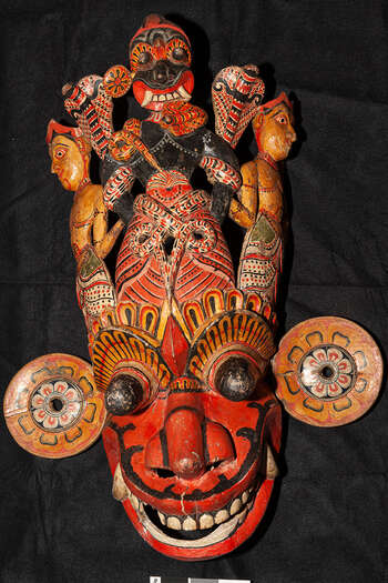 Rituell funksjon og kontekst: Kōlam-teater / Bali / Garāyakumā UEM7001 representerer en djevelfigur fra Rakshā-kulten. Masken kan representere enten «Fyrsten av Støttannen» ( rakshā dala-kumarā ) eller Anāňga-yakkā, også kjent som Anāňga-bahirawa eller Watā Kumara. Karakteren kan også ha blitt brukt som en maske for Kalū kumāra, et overnaturlig vesen som gjør unge kvinner syke. Et annet ritual som masken kan ha blitt brukt i, er Garāyakumā, for å representere Dalā-Kumārā, en av inkarnasjonene av Garā Yakā. De viktigste ikonografiske trekkene ved UEM7001 er: • Flere figurer på toppen av masken som er relatert til maskens hovedansikt • Sirkulære øreornamenter festet separat som symboliserer solen og månen • Øreornamentene er dekorert med eldre Sinhālese-motiver: en blomst med åtte kronblader ( atā-pethi -mal-mosthara ) som knytter masken til Garā-yakā. • Den stående figuren i svart på toppen av masken kan representere både karakterene Garā-yakā eller Kalū kumarā (demon for seksuell lyst) • På siden er to unge guddommer, trolig mann i forbindelse med Garā-yakā, eller kvinne når masken er av Kalū kumarā eller Anāňga-Yakkā Merk: Den røde huden kan også referere til karakteren Riri Yaǩkā eller en av hans inkarnasjoner. Tradisjon: Bali/Sanniyakumā/Shānthî, Kārmā/Tōvil/Kōlam-teater/sørlig dansetradisjon/lavlandet. Denne tradisjonen er populær i Mātara, Gall, Bentōta og Ambalaňgoda i Sørprovinsen og i Olabodūwa, Rāiygama Kōralē, i Vestprovinsen på Sri Lanka. Materialer og teknikker: Polykromt utskåret tre (hvitt, gult, rødt, blått, grønt og svart).
